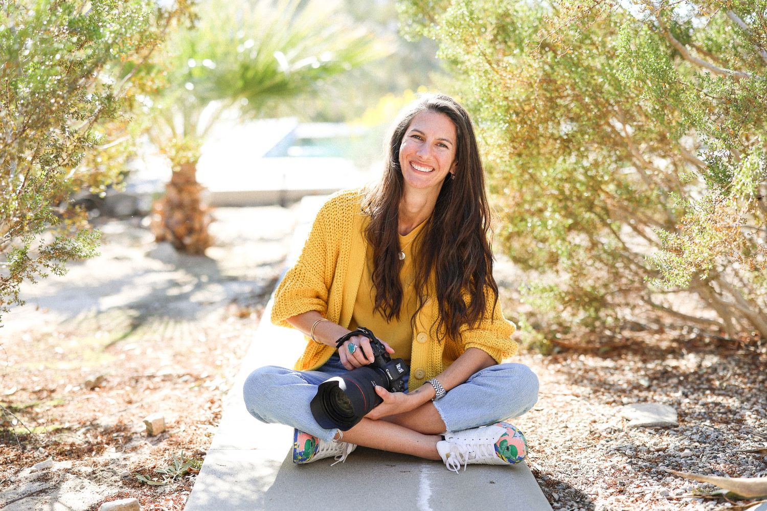 Woman in yellow sweater sitting cross-legged outdoors with bright natural lighting.