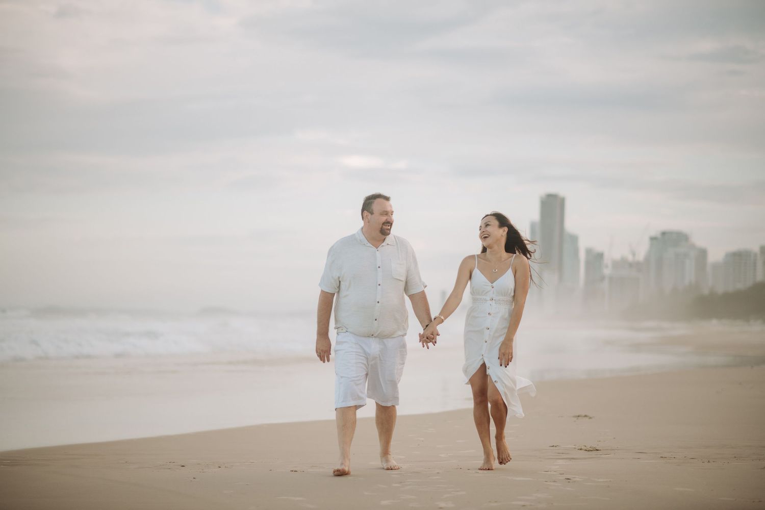 A couple walks hand in hand along a misty beach with a city skyline in the background.
