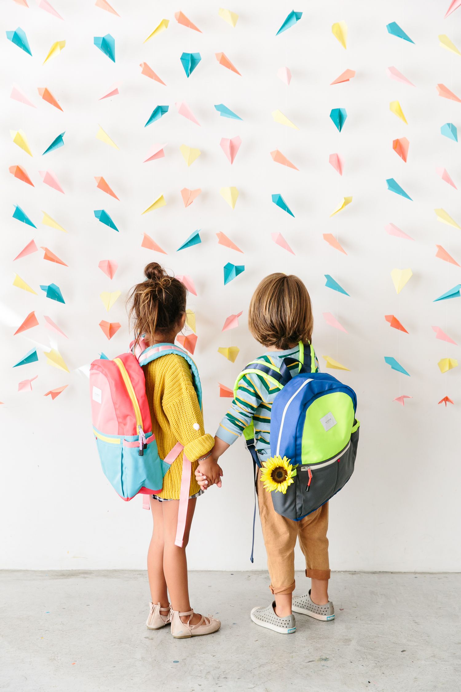 Two young students wearing colorful backpacks stand against a wall decorated with pastel paper shapes.
