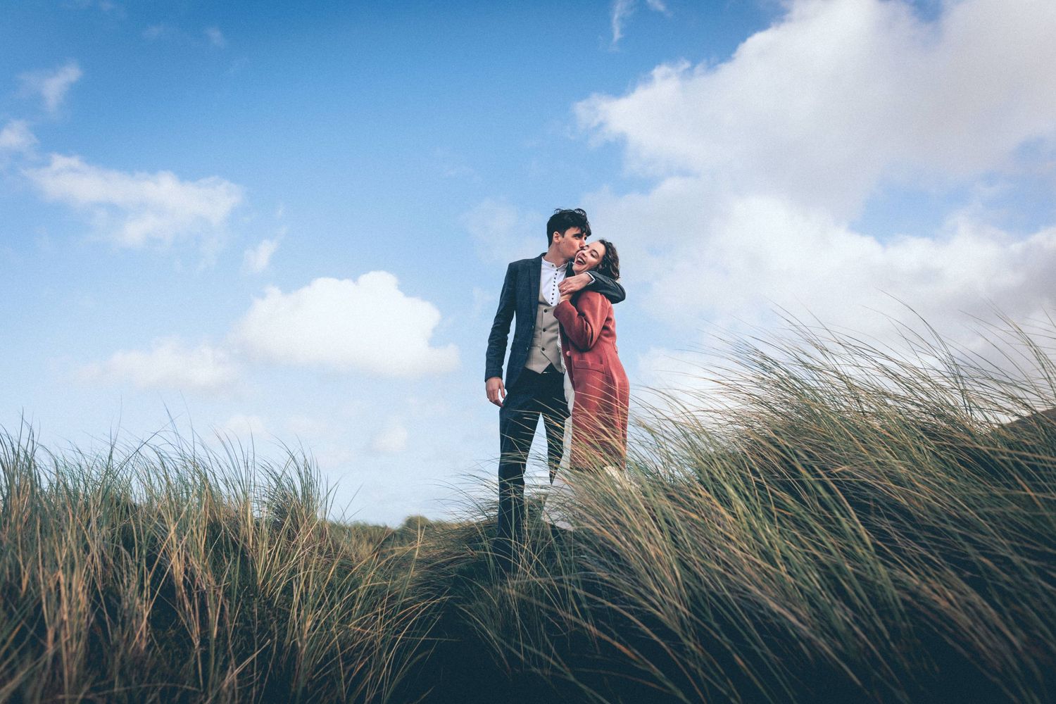 A couple stands together in tall swaying beach grass against a blue sky on a bright sunny day.