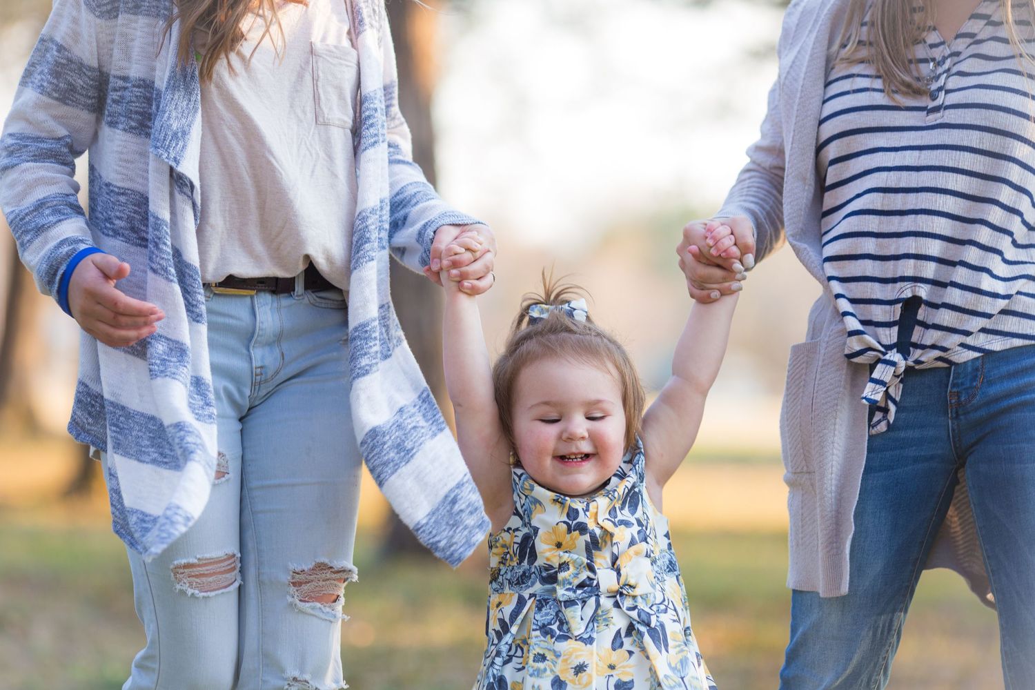 Two women holding hands with a happy toddler girl in a floral dress outdoors.