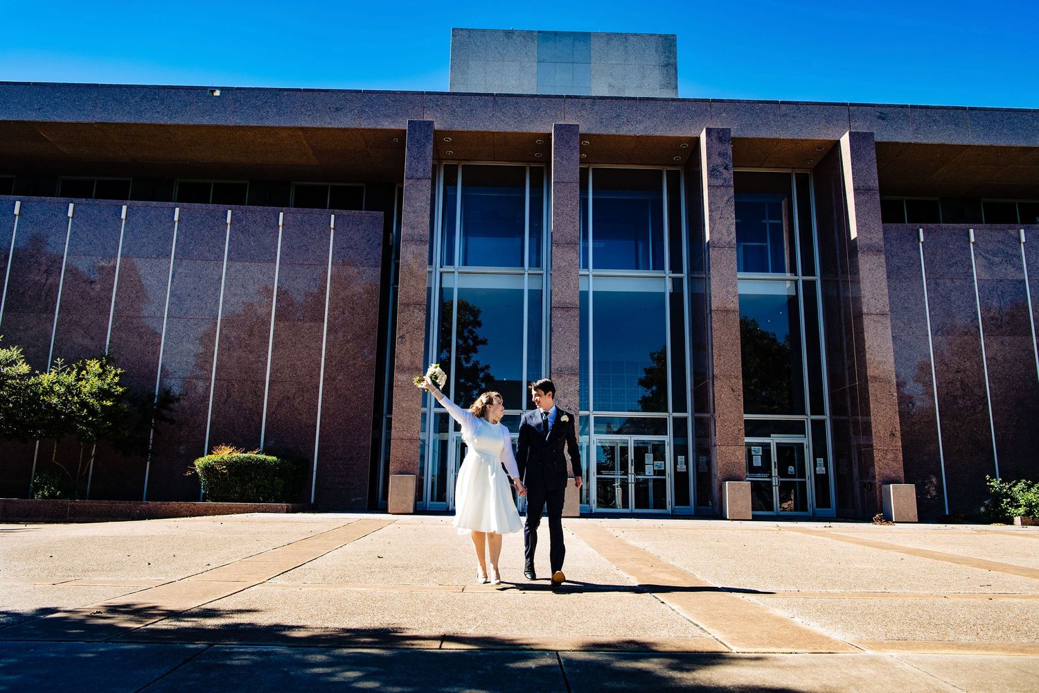 A couple celebrates outside a modern building with glass doors on a sunny day.