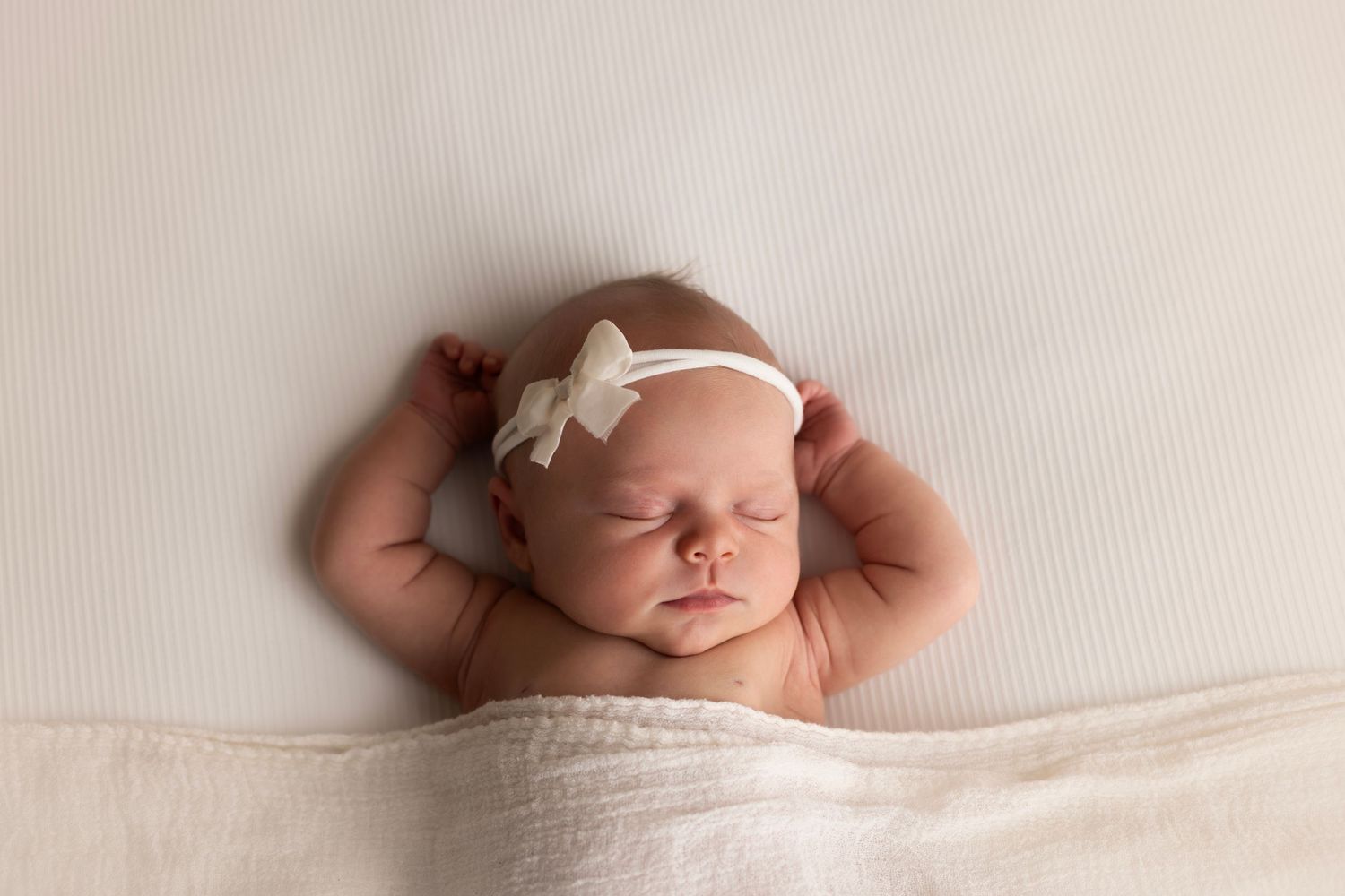 Newborn baby girl laying on a white blanket with a white headband, sleeping with her arms above her head.
