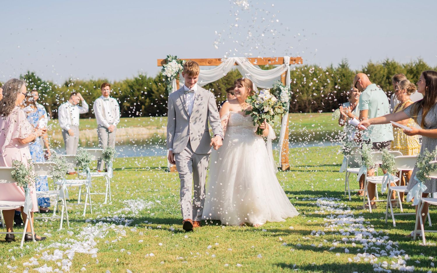 Bride and groom walking down outdoor aisle as guests toss flower petals at sunny garden wedding.