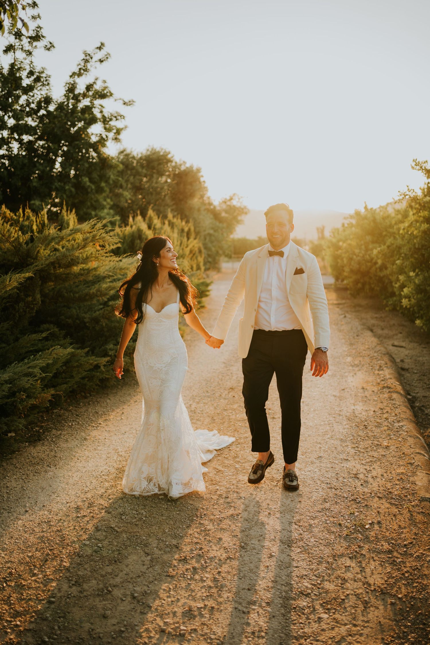 Romantic sunset photo of newlyweds walking hand in hand on a dirt path.