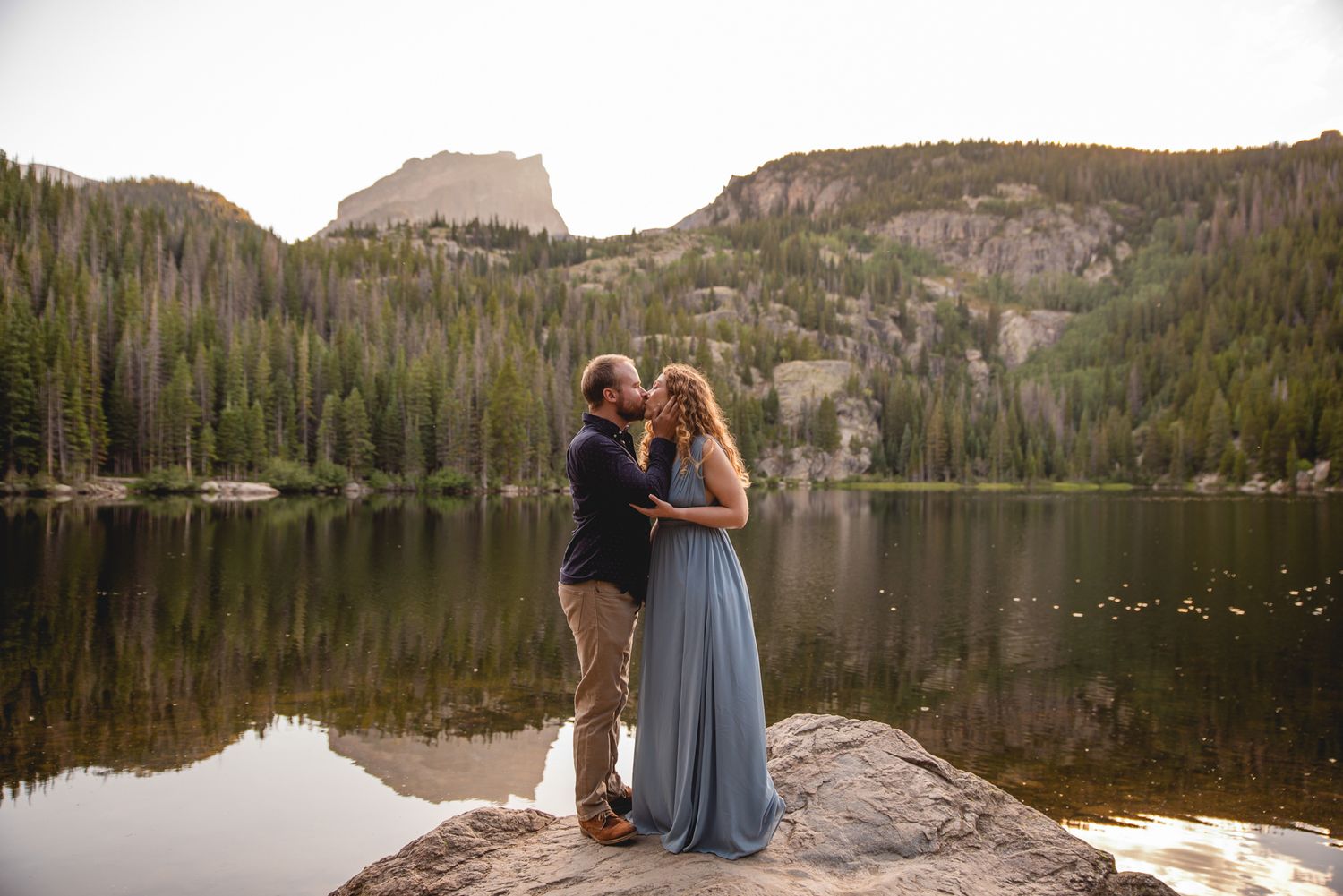 A couple embraces while standing on a rock overlooking a serene mountain lake with evergreen forest views.