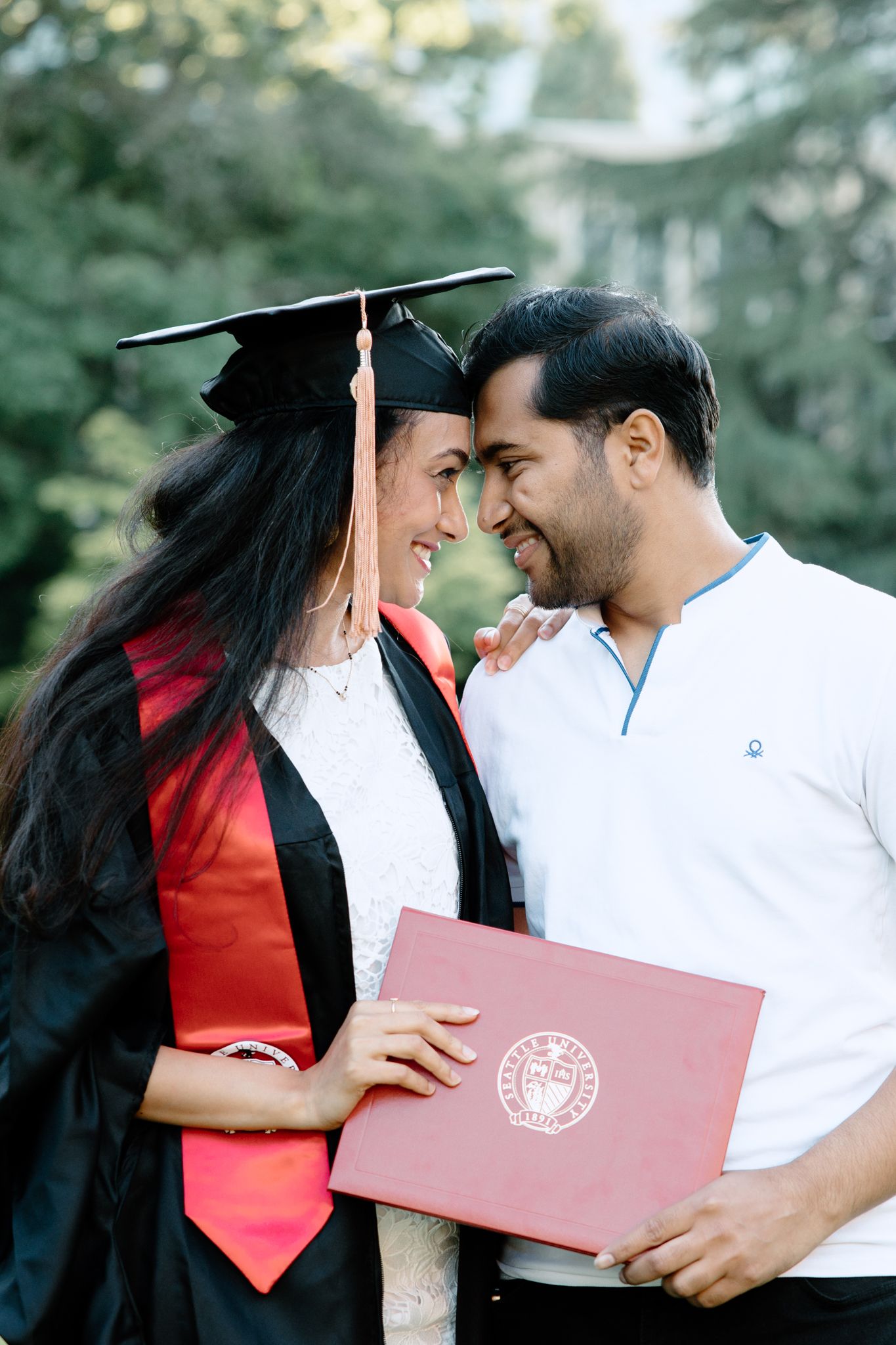 Graduation celebration moment with a graduate in cap and gown holding a red diploma folder against a natural background.