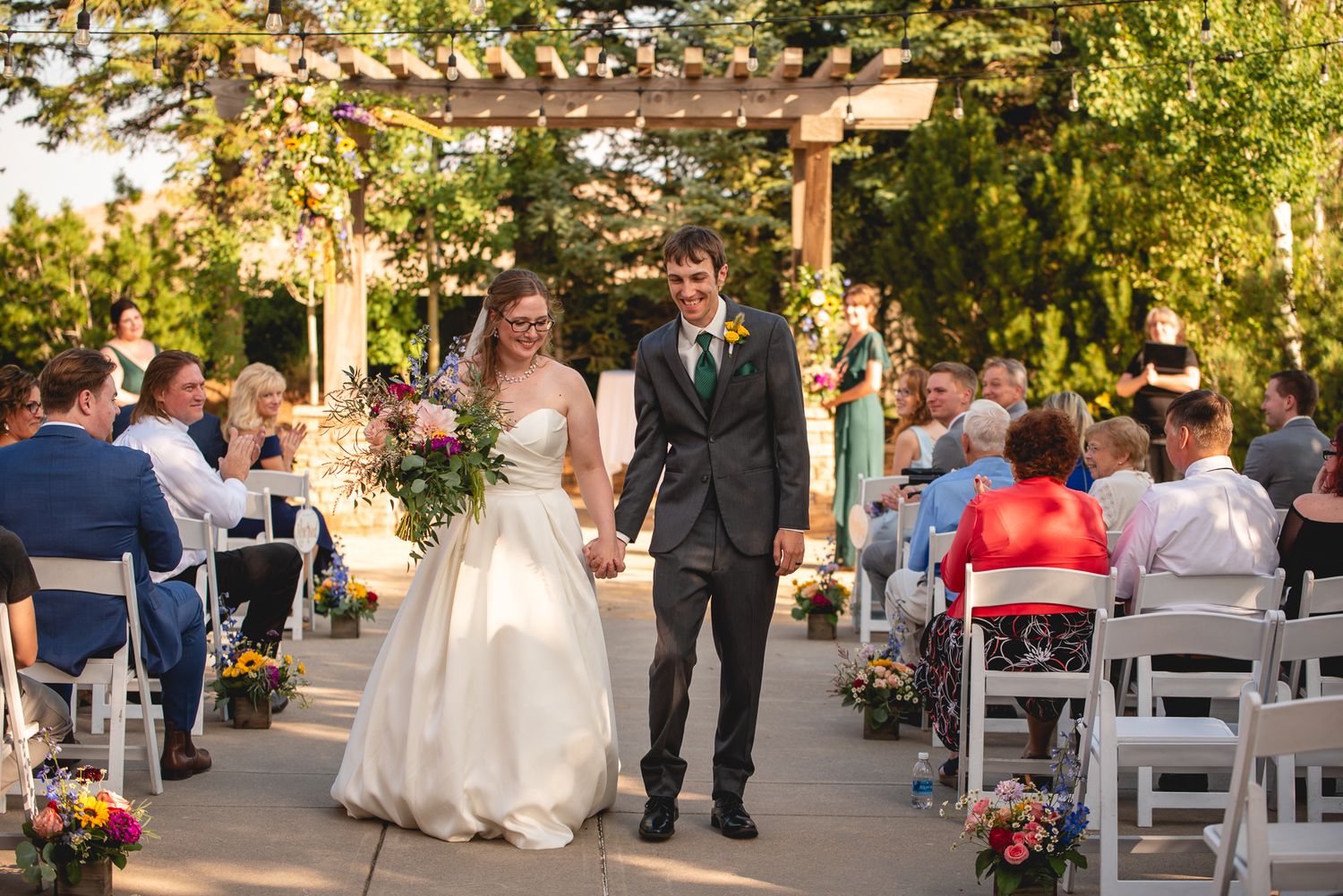 A couple exits their outdoor garden wedding ceremony under a wooden pergola with floral decorations.