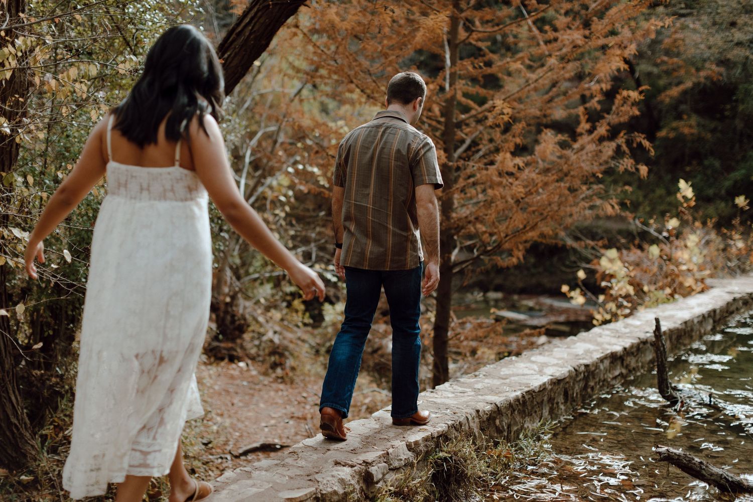 A couple walks along a creek path during golden hour, with the person in a white dress holding hands with their partner.