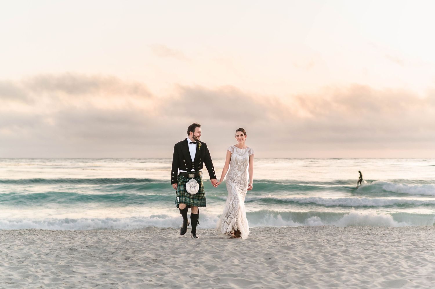 A couple strolls on a beach at sunset, with one wearing a traditional Scottish kilt.