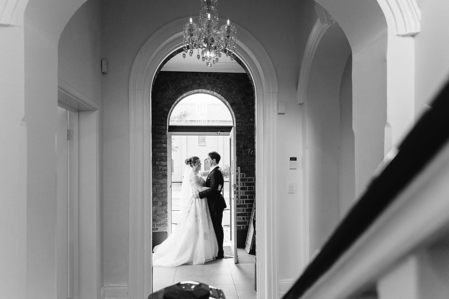 A silhouetted wedding couple stands in an elegant white archway with chandelier above.