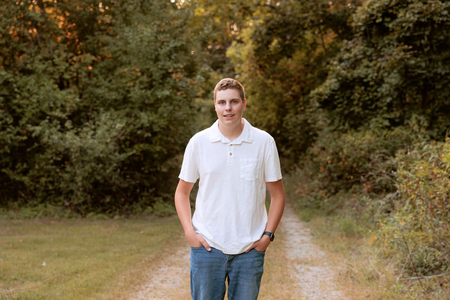 Casual outdoor portrait in white polo shirt standing on dirt path surrounded by greenery.
