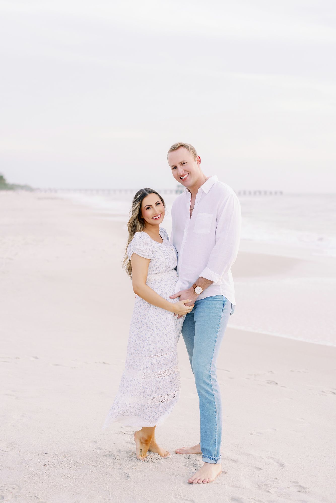 Happy couple in casual white outfits poses together on a beach at sunset.