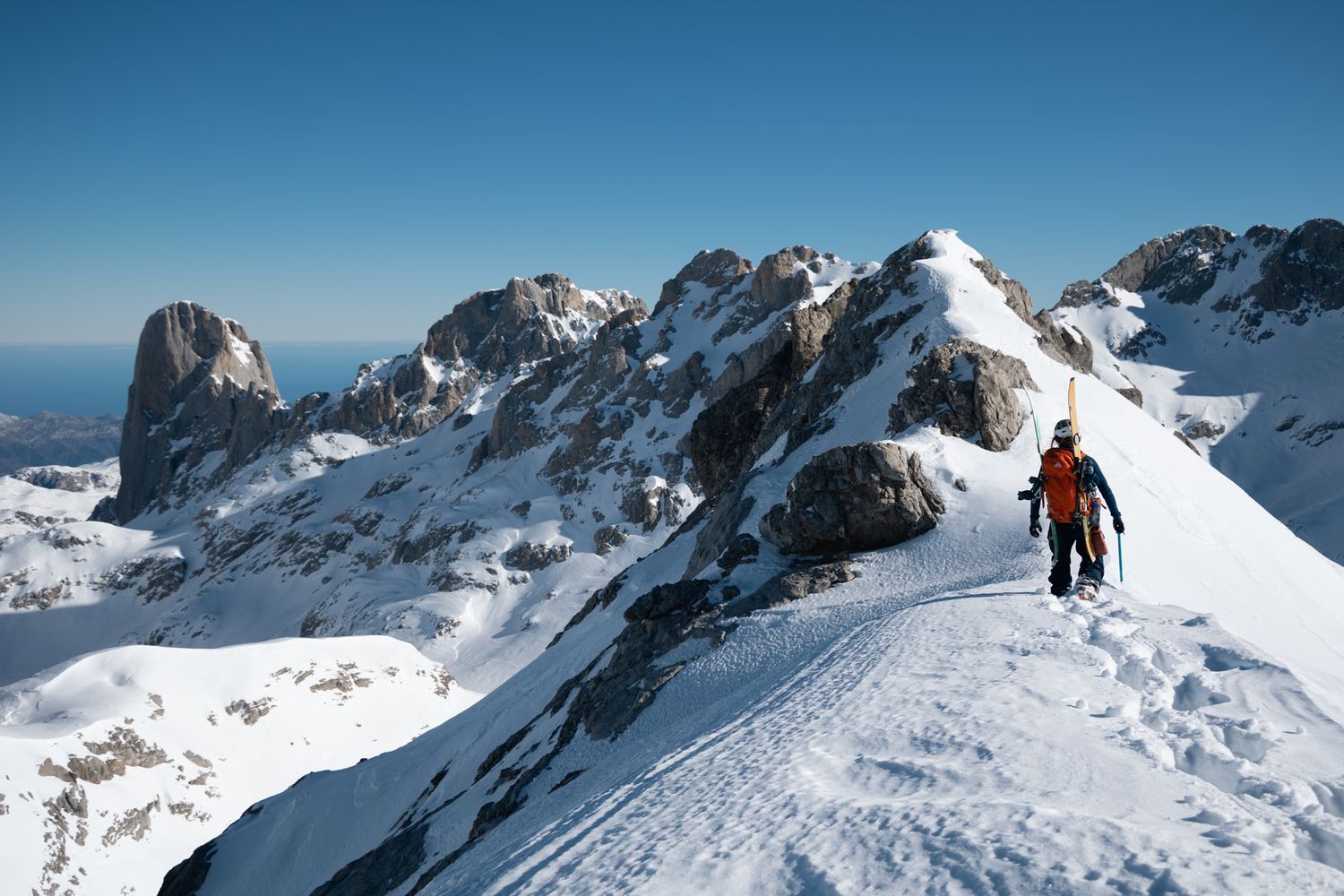 Woman with orange backpack walks on a snowy ridge toward a peak in the sunshine, mountains and ocean in the background.