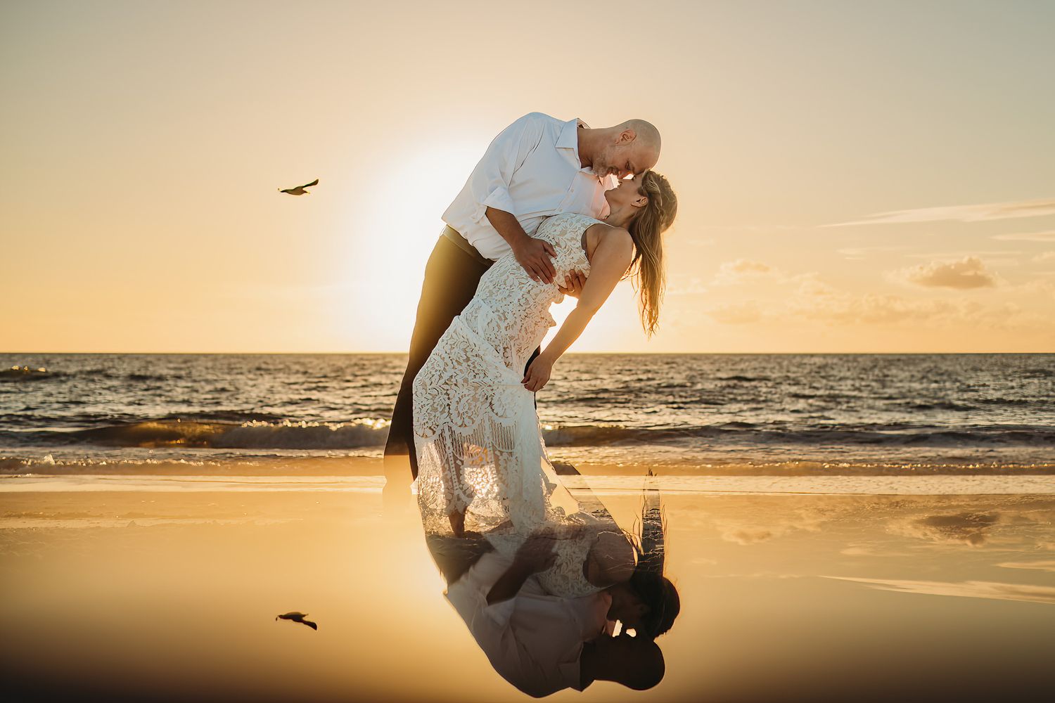 A couple dances and plays on the beach at sunset with seabirds flying overhead.