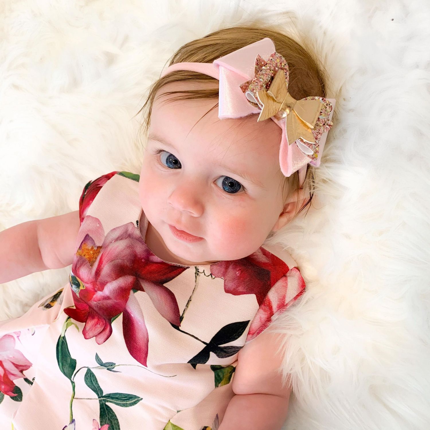 Baby in floral dress wearing pink and gold bow headband on white furry backdrop.
