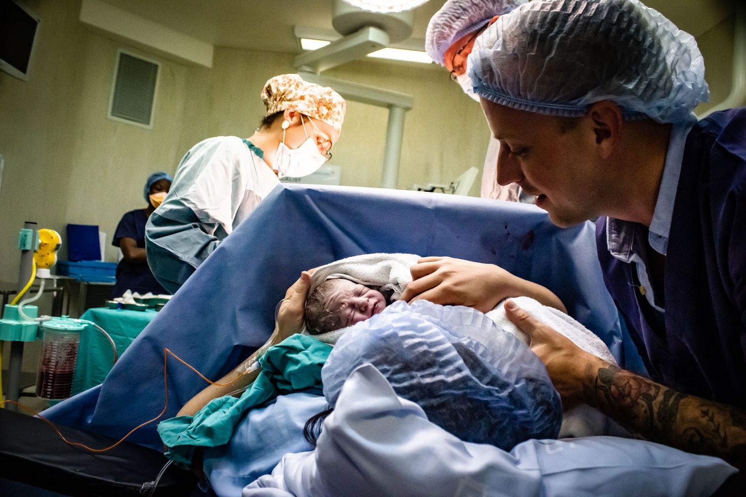 father and mother holds newborn baby in theatre doctor working in background blue hair net