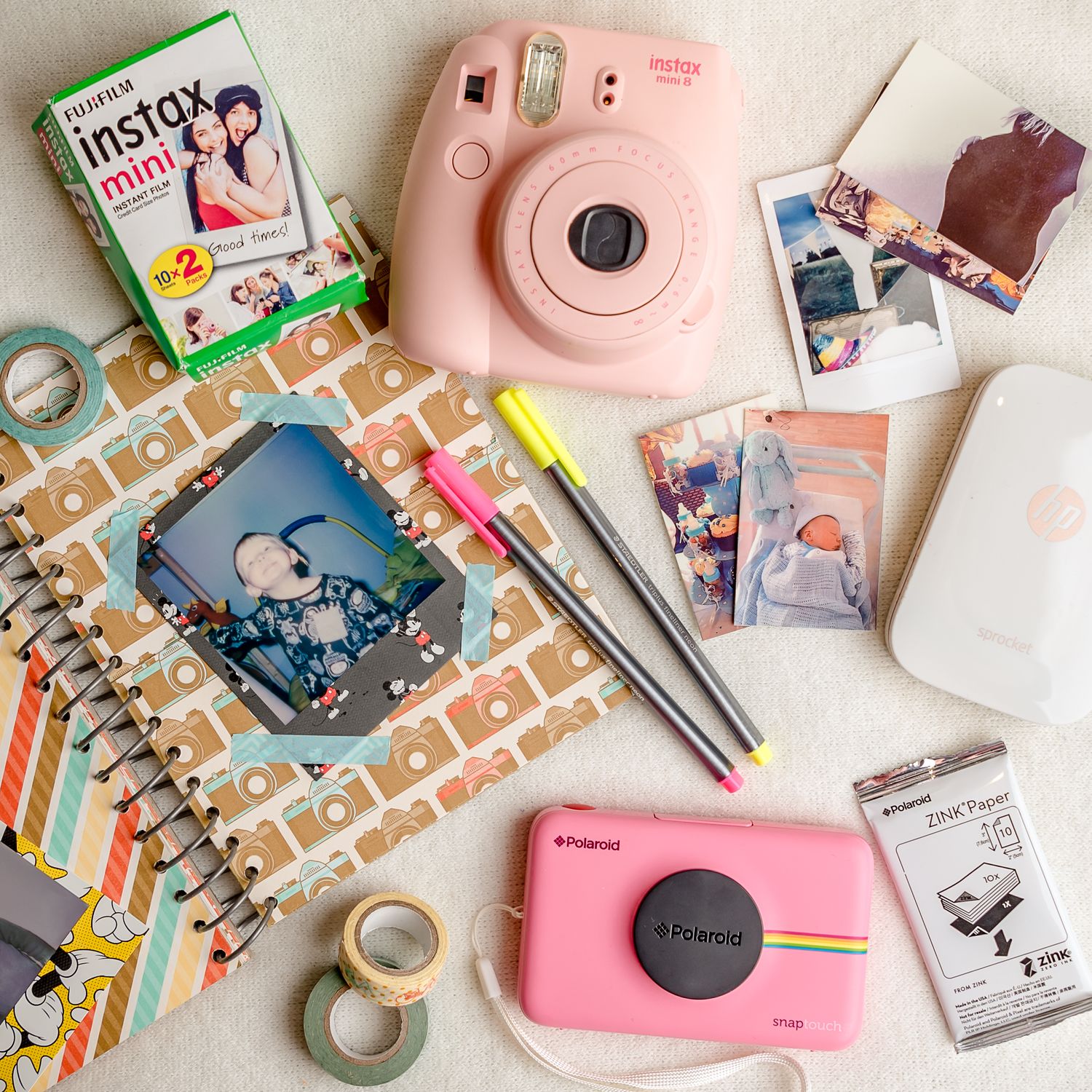 Pink Instax camera with photo prints, washi tape, and stationery supplies arranged on a white surface.