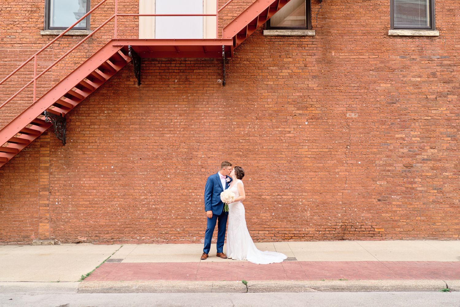 A romantic wedding photo against a rustic brick wall with red fire escape stairs.