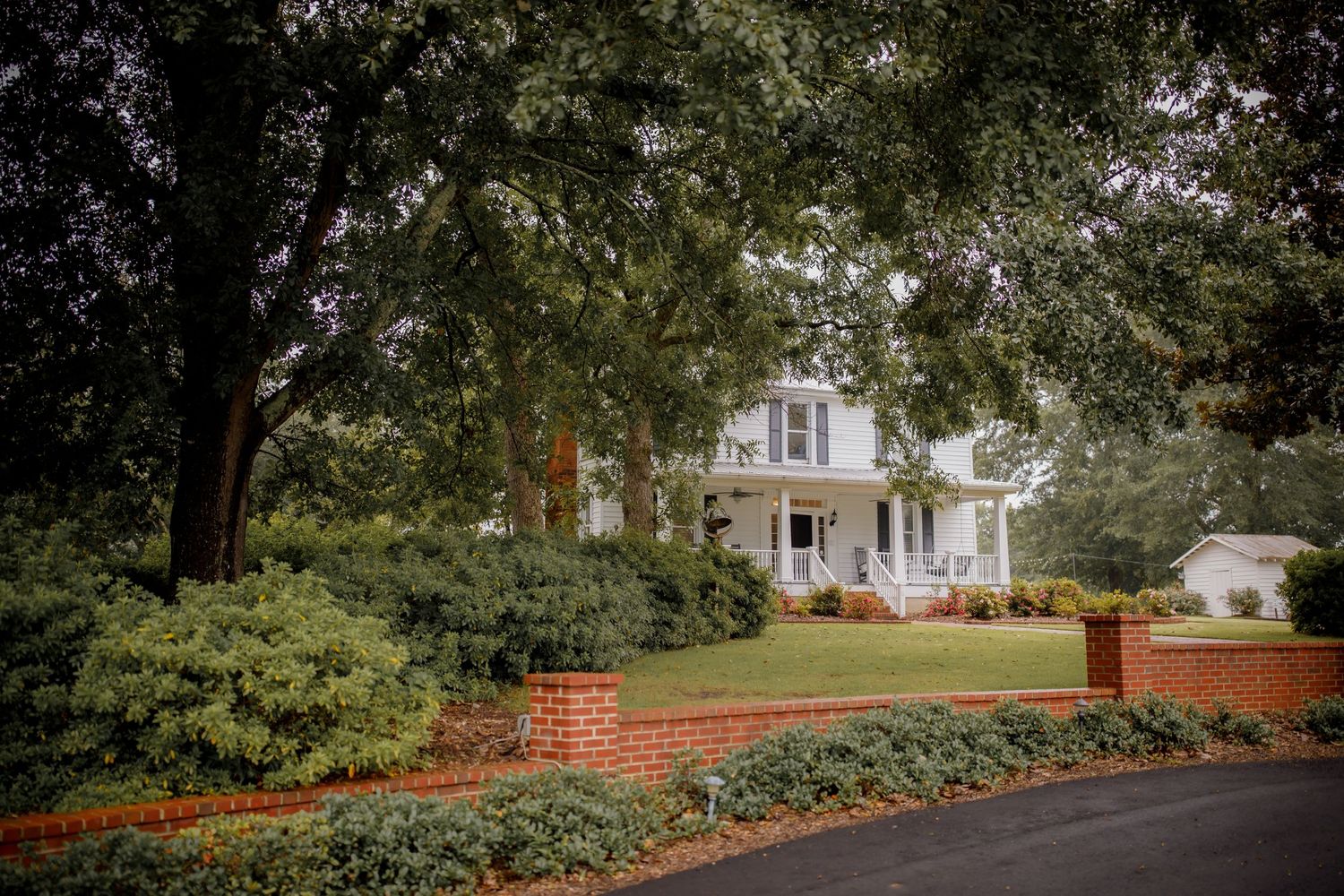 Classic white Southern house with brick wall and lush landscaping viewed through tree branches.