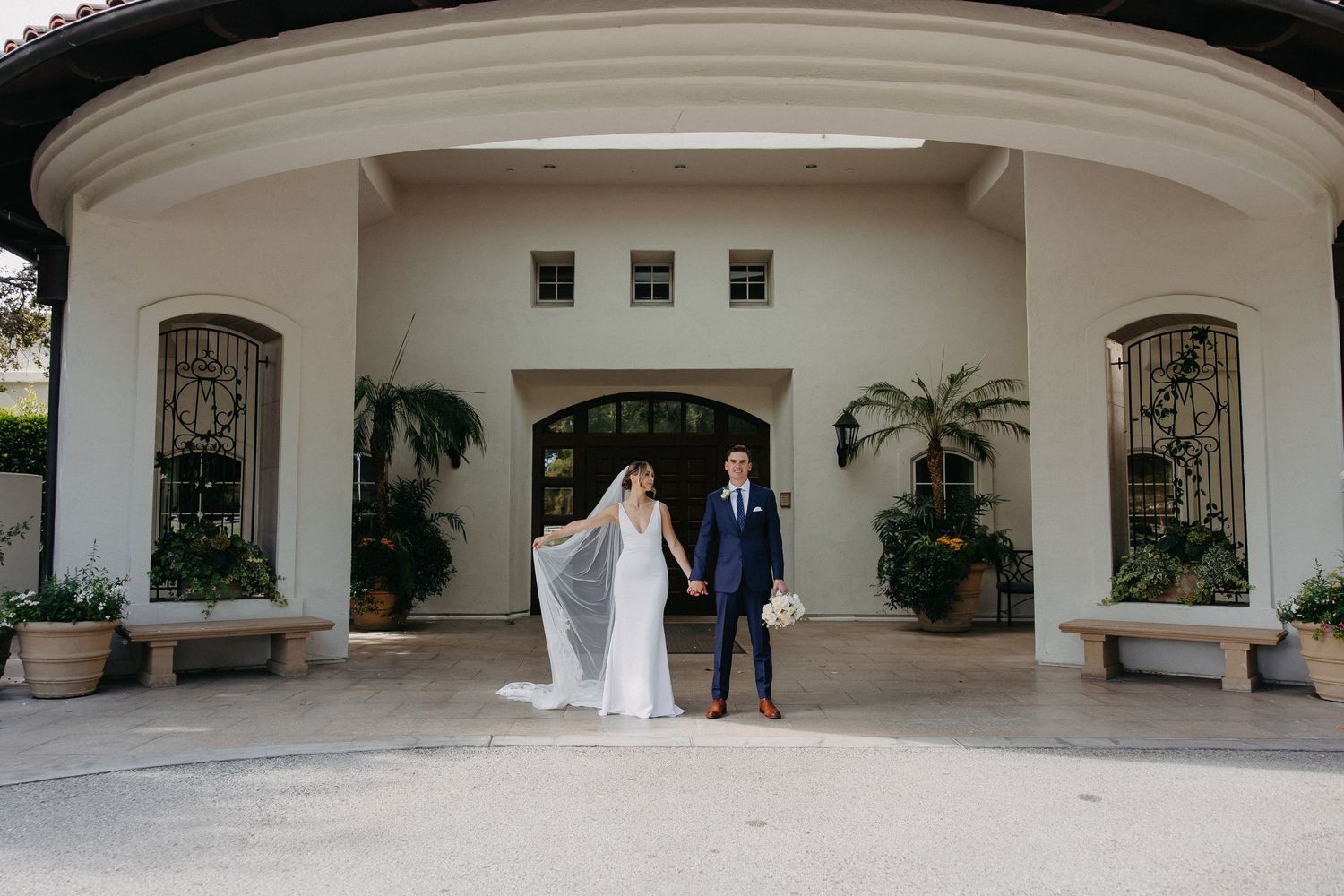 Wedding couple exits elegant white archway entrance with tropical plants and benches.