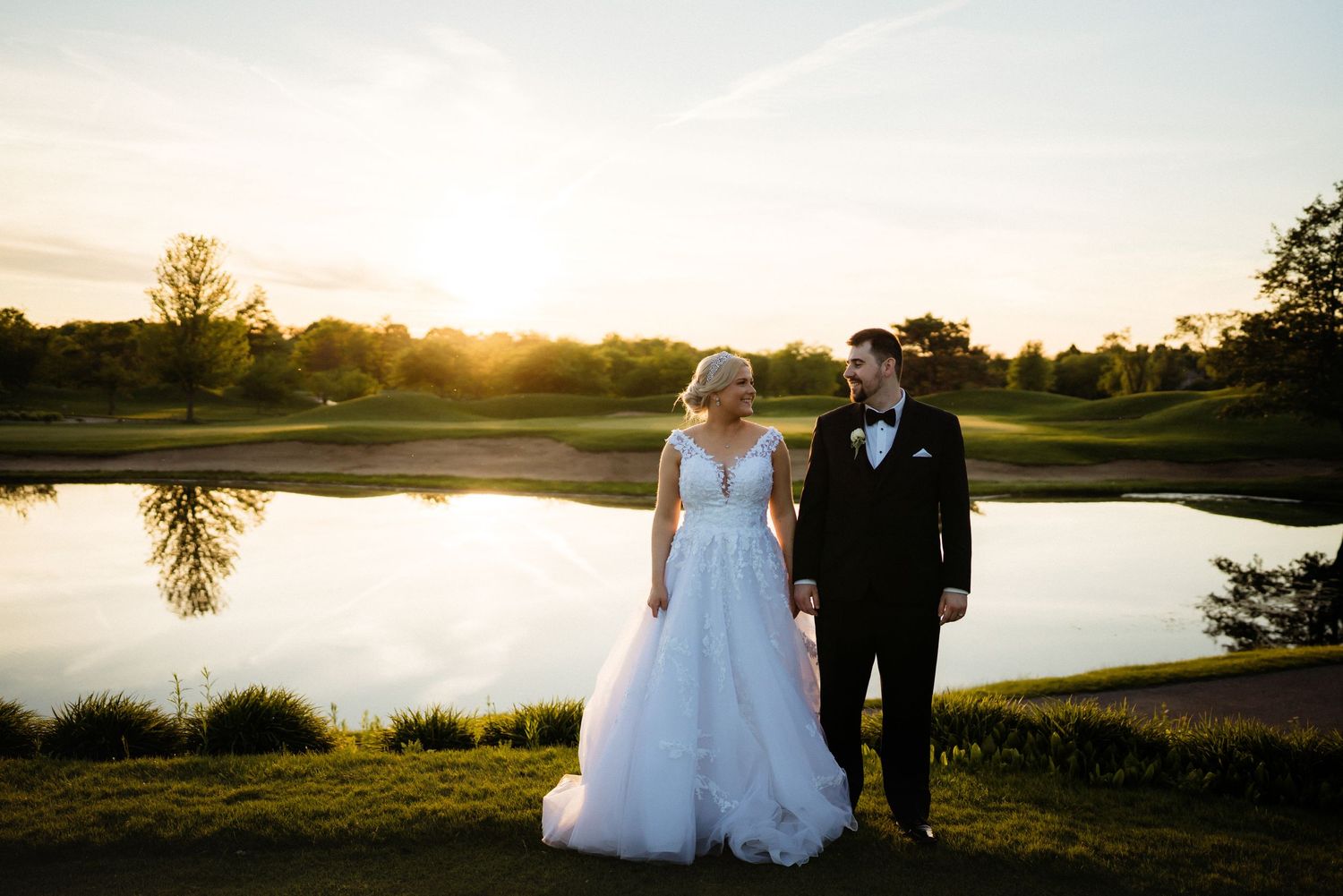 A couple stands together at sunset by a tranquil pond in formal wedding attire.