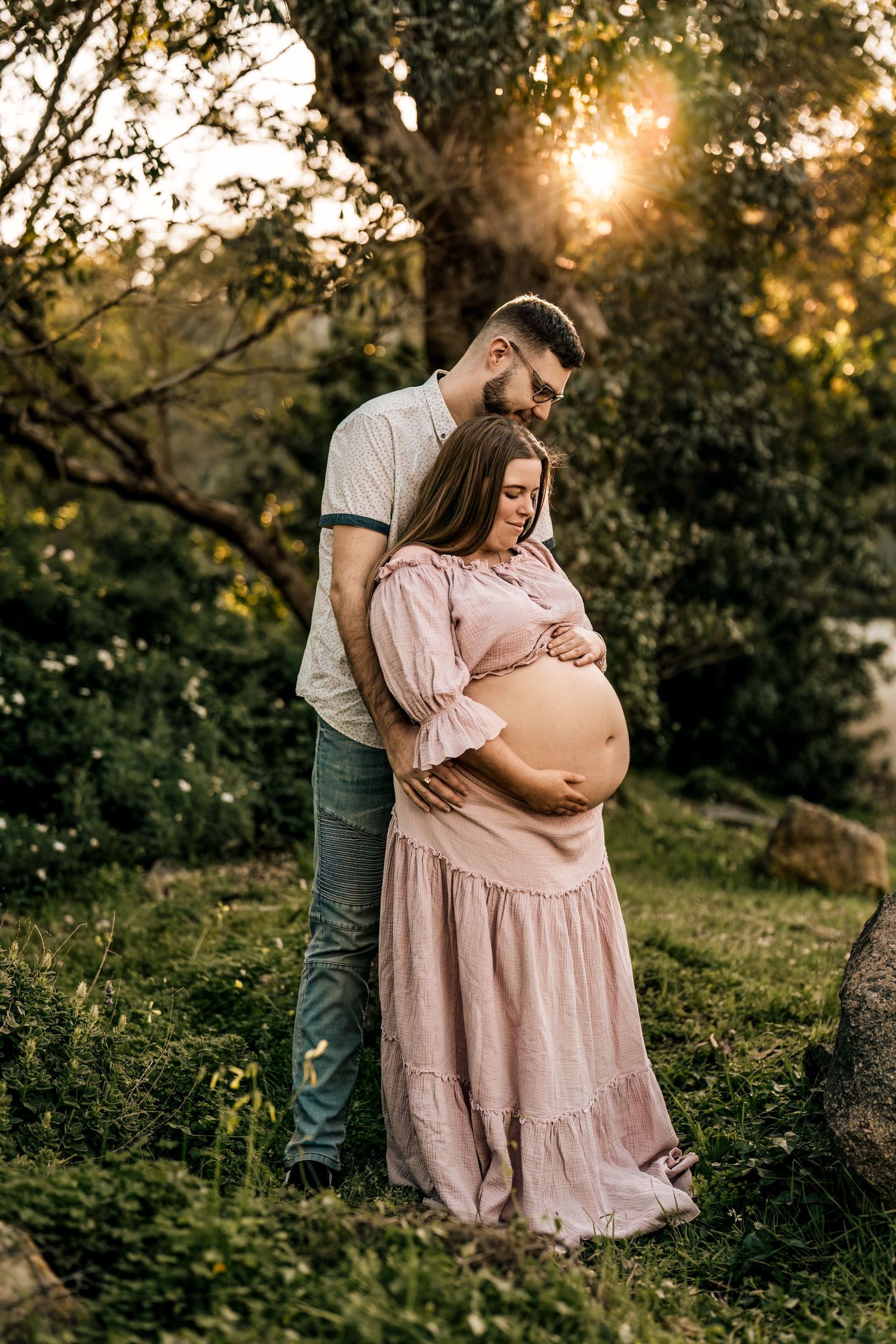 Intimate maternity photo of couple embracing outdoors at sunset with woman in pink dress.