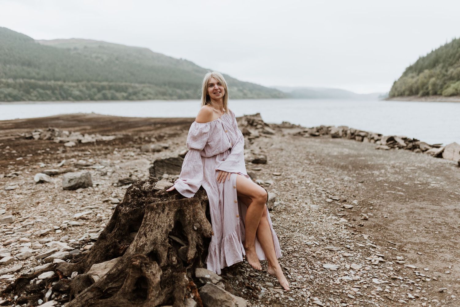 A person in a flowing mauve dress poses on a tree stump by a misty lake surrounded by mountains.