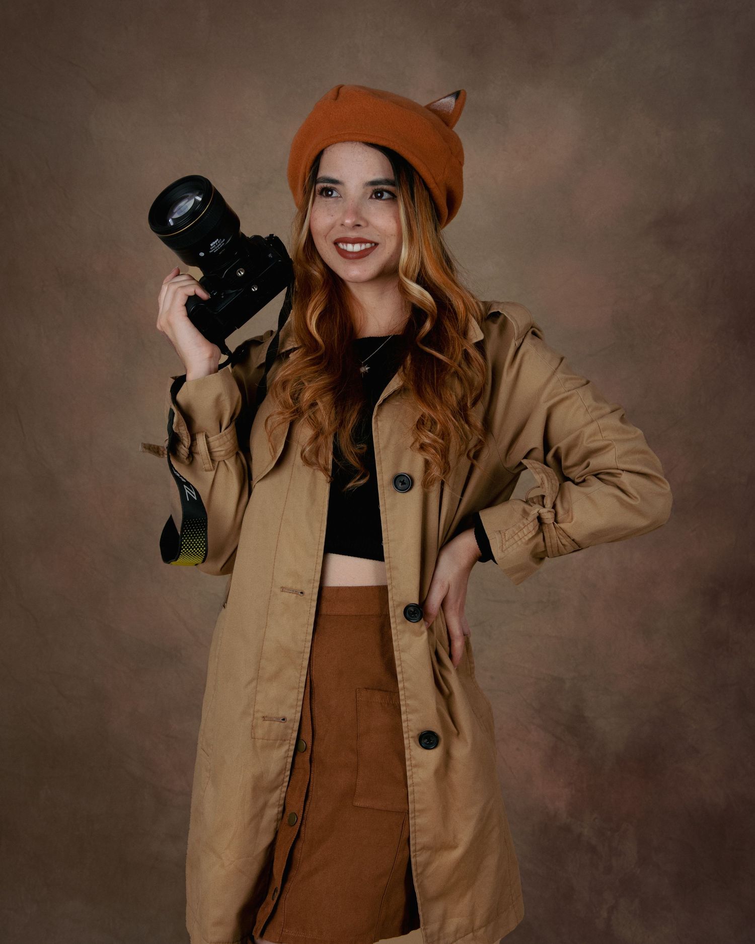 Photographer in fox hat and tan coat holds camera in professional portrait setting.