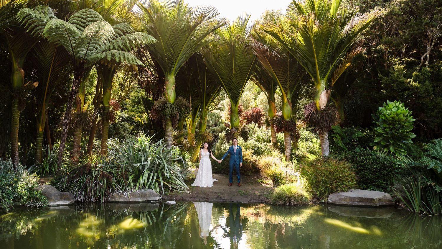 A scenic wedding photo reflected in a tranquil pond surrounded by tropical palm trees.