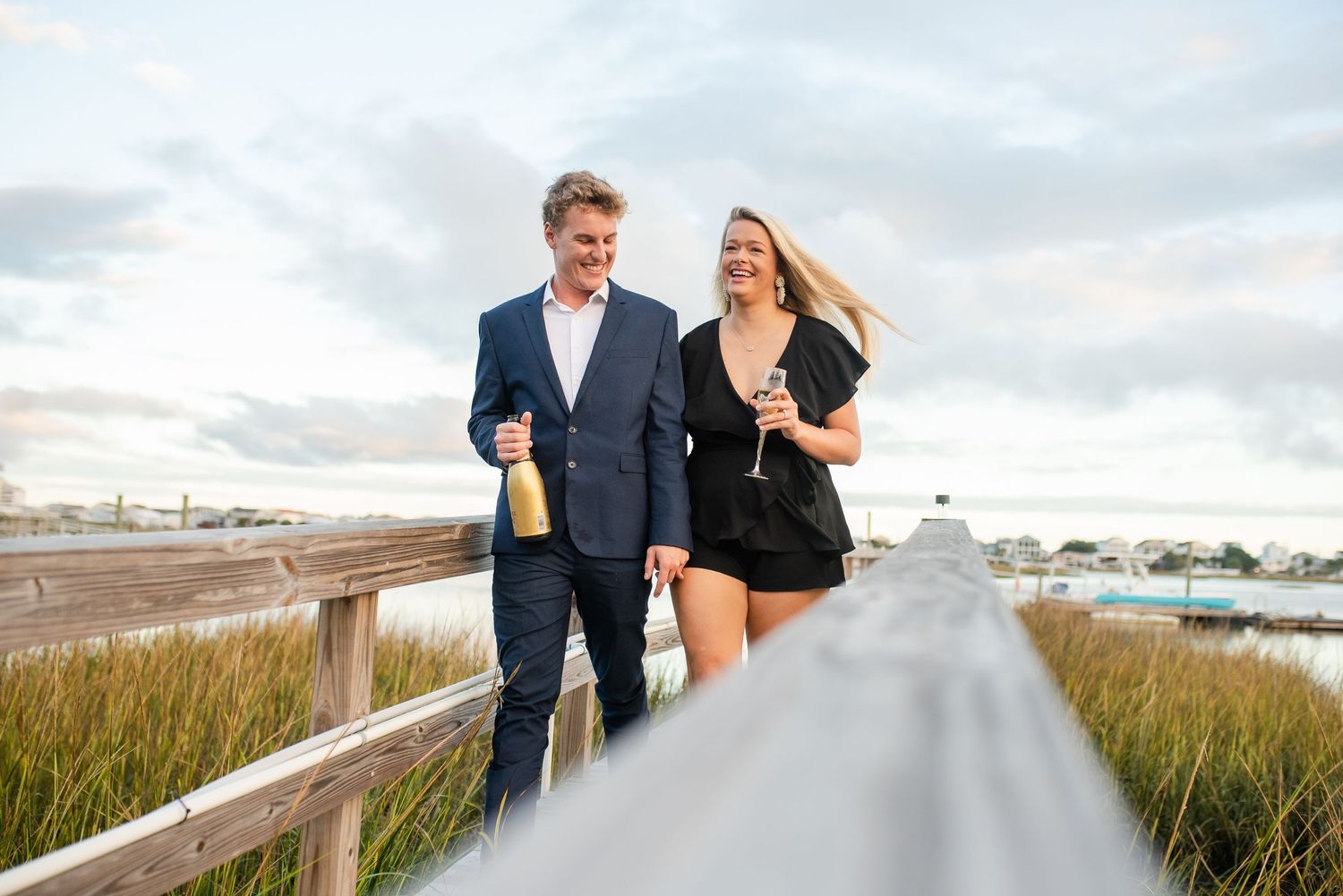 A couple walking on a boardwalk through marsh grass at sunset.