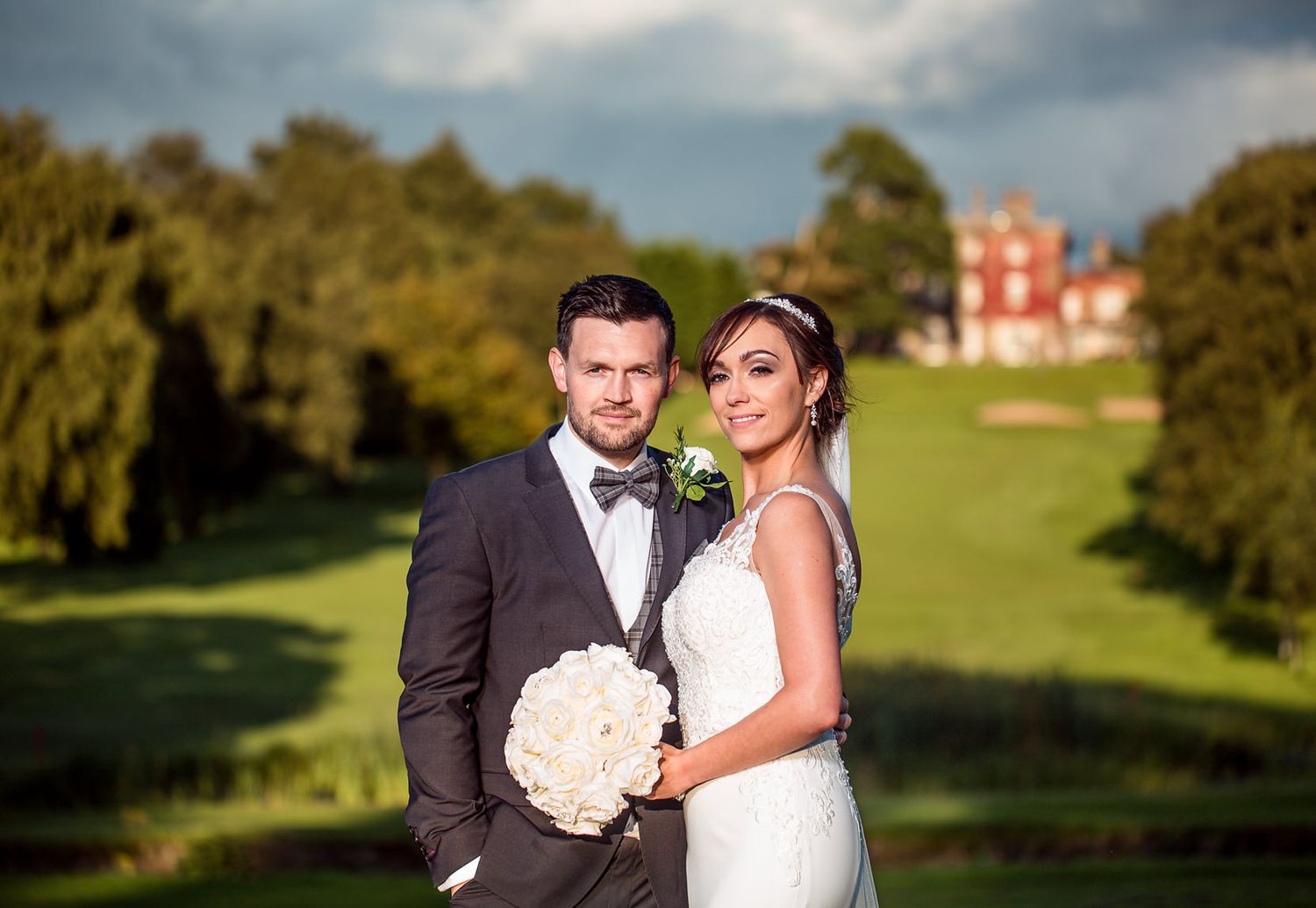 A wedding couple poses outdoors with a grand estate building visible in the background.
