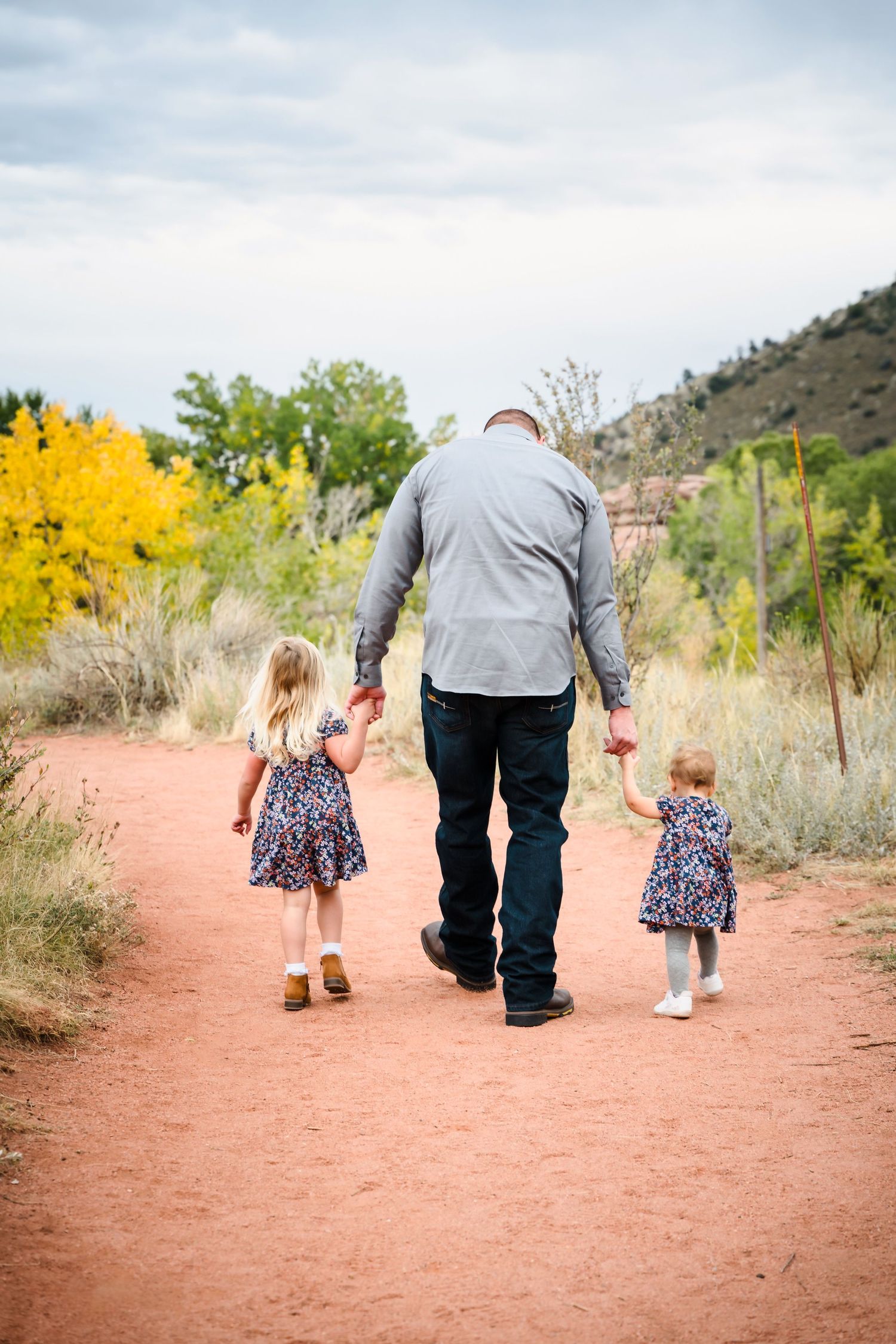Adult walking with two young children on a red dirt trail surrounded by desert landscape and autumn foliage.