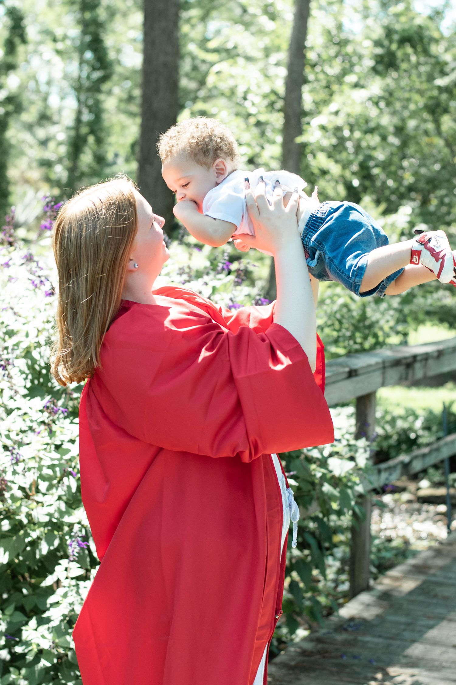 Graduate in red gown lifts joyful toddler in blue pants during outdoor celebration.