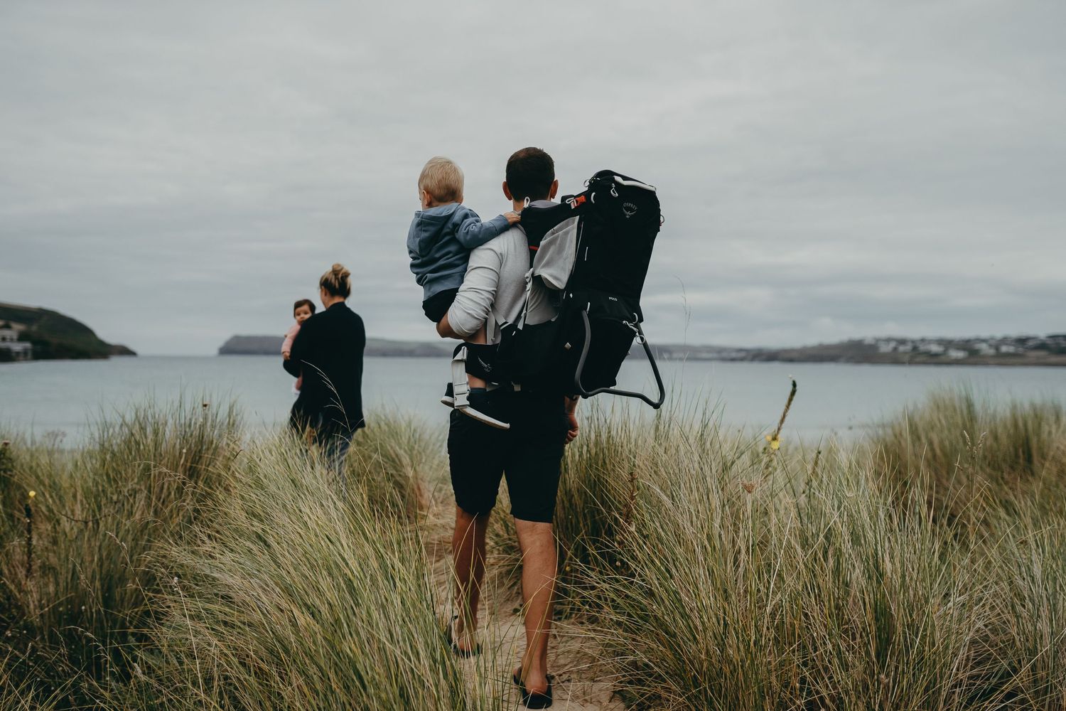 Figure carrying child and backpack through coastal dunes with ocean view.