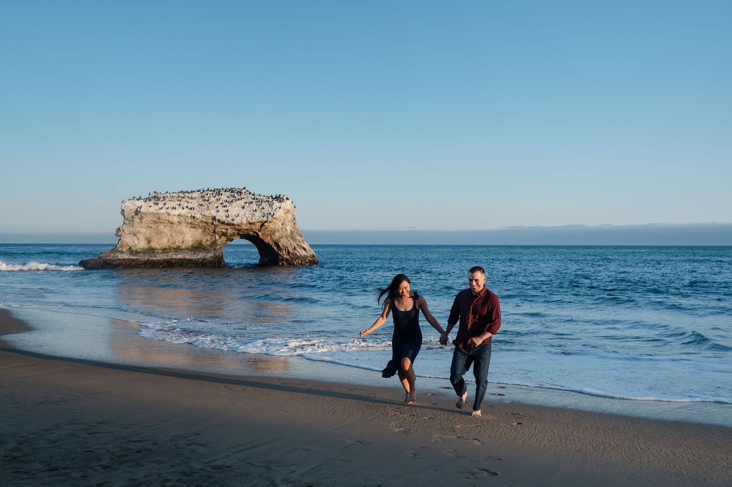 A couple runs joyfully along the beach with a natural rock arch formation in the background.