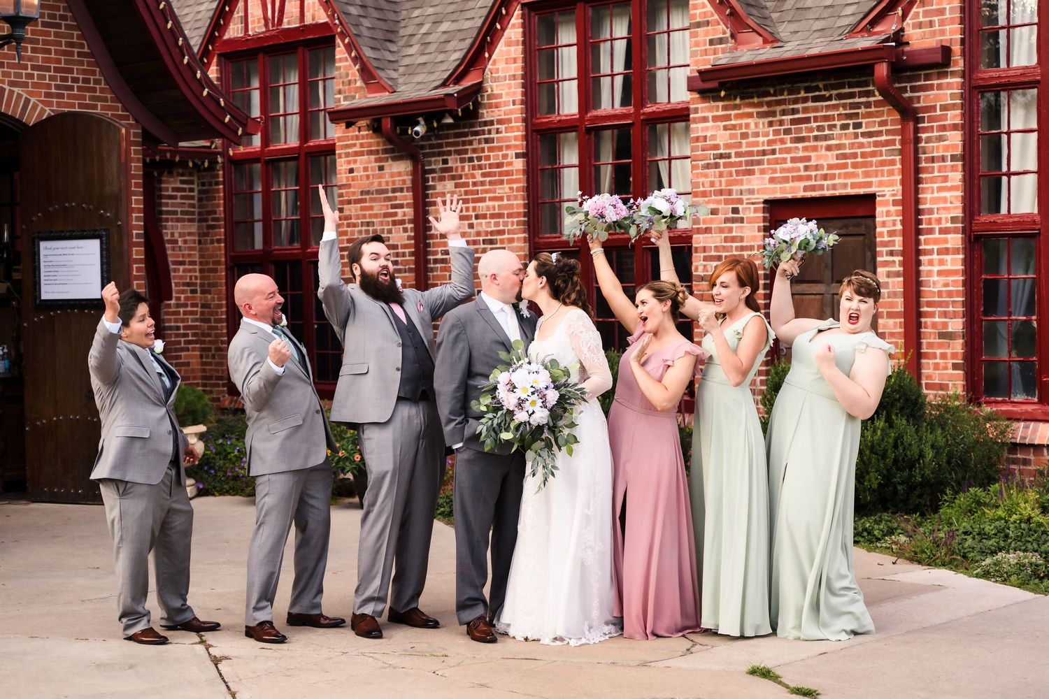 Wedding party celebrates outside a brick building with bridesmaids in pastel dresses and groomsmen in gray suits.