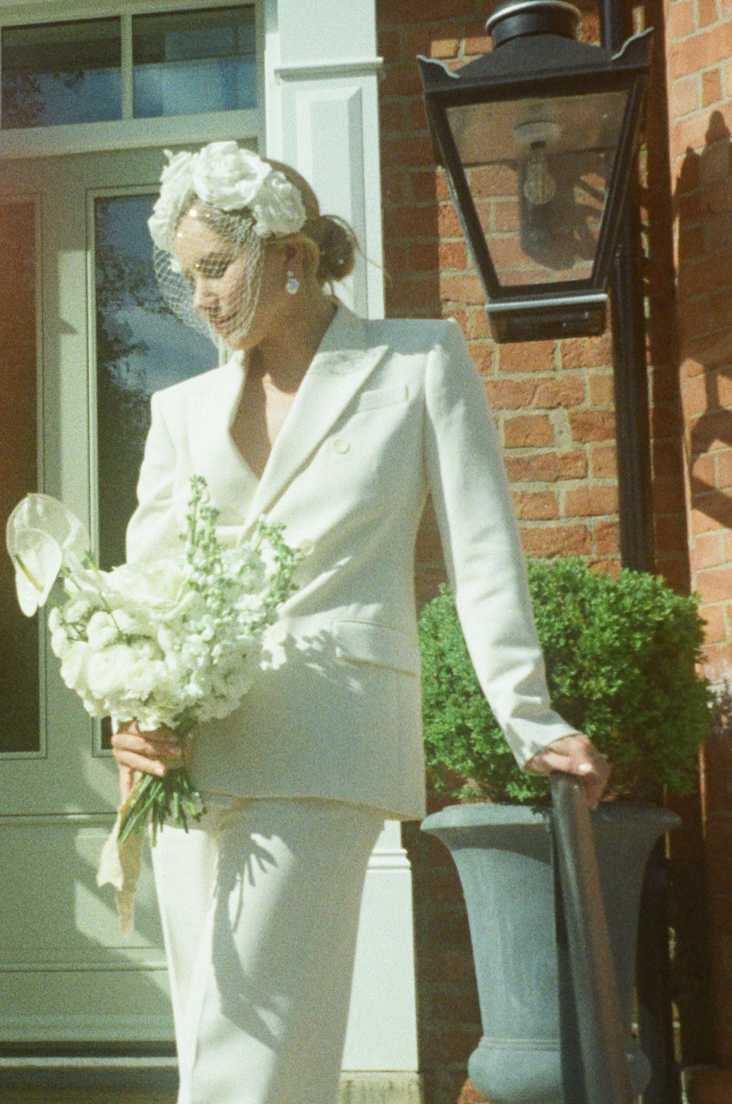 A figure in an all-white ensemble holding a bouquet near a brick building with a vintage lamp.