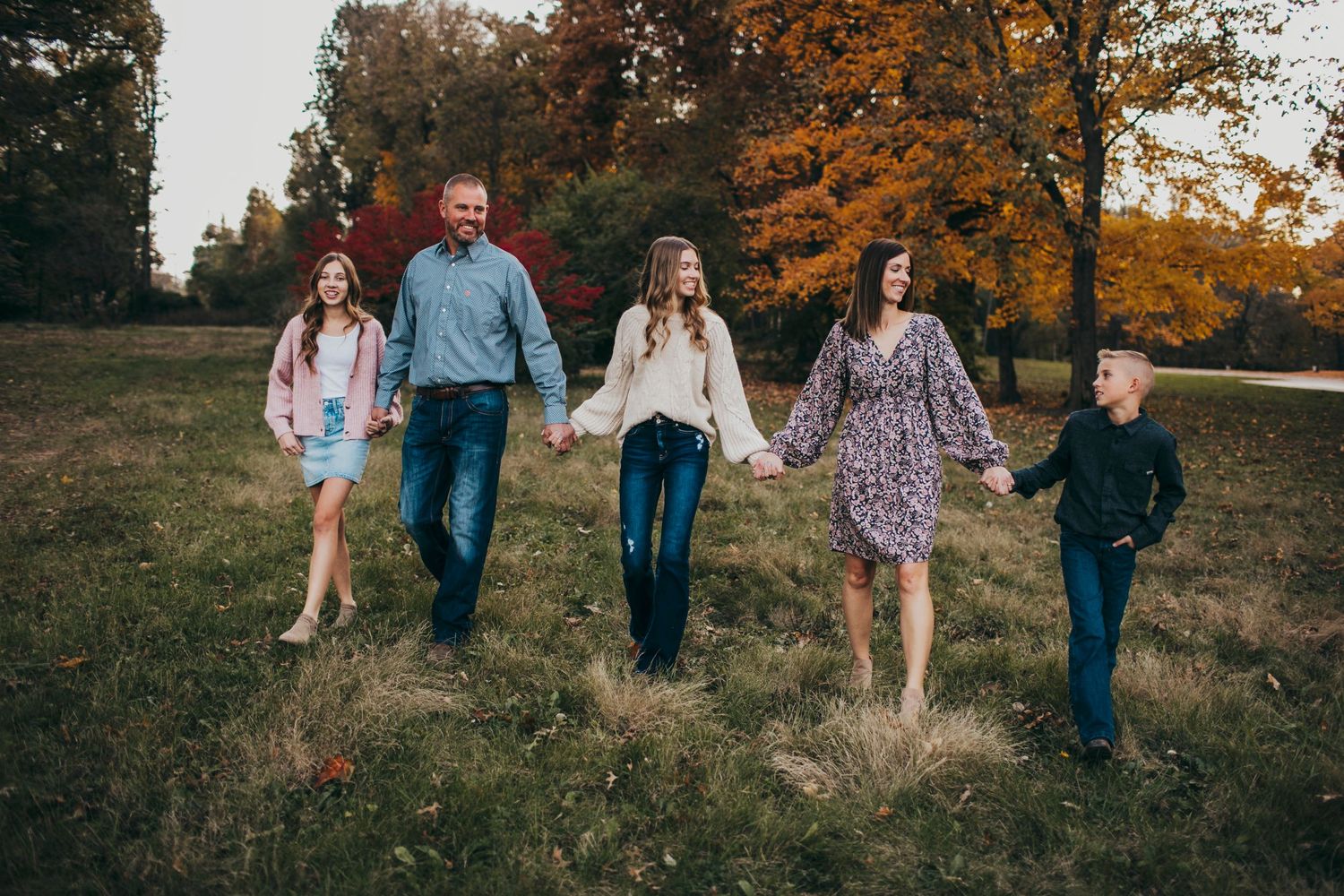 Family of five walking hand-in-hand through an autumn park with vibrant orange foliage.