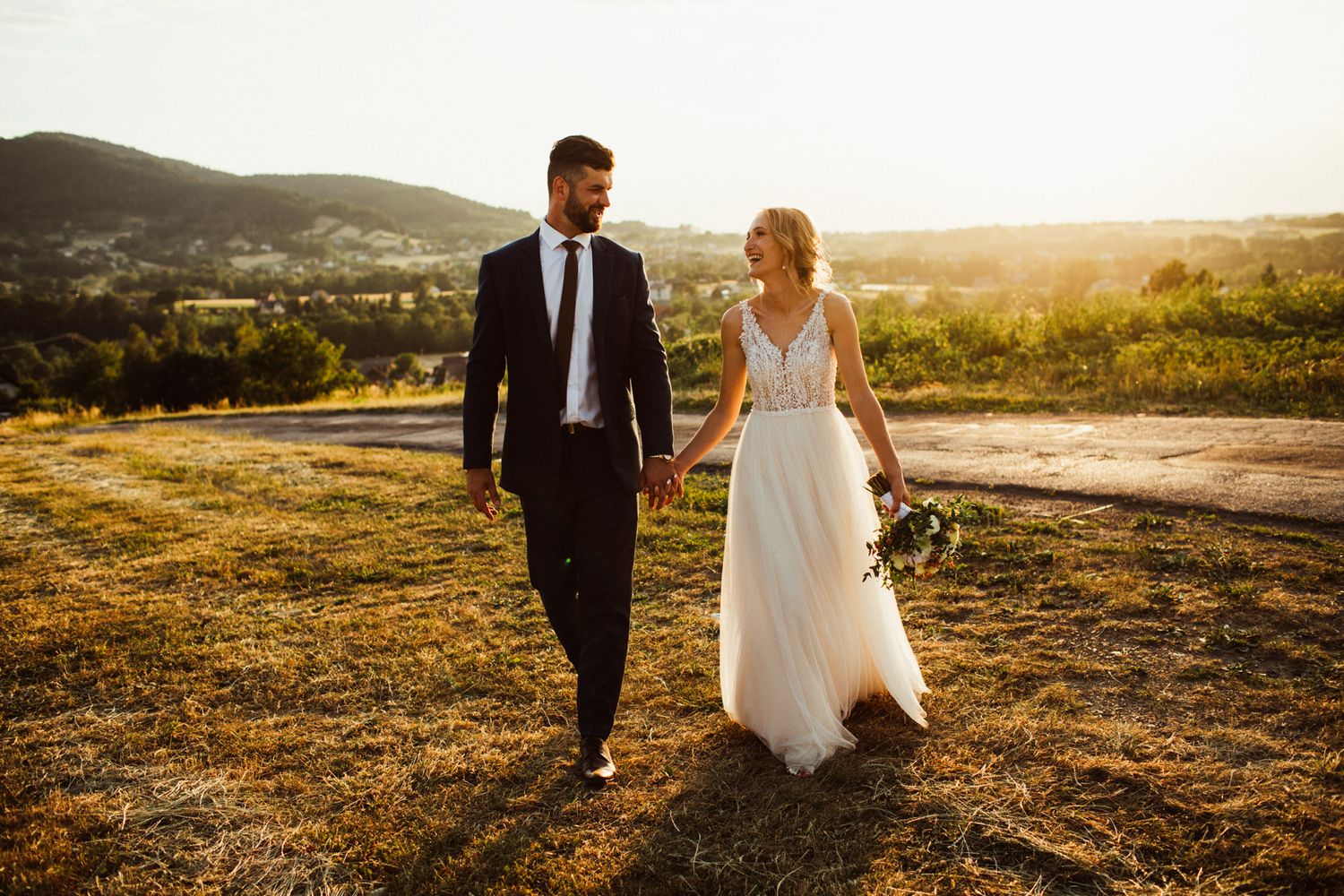 Bride and groom walk hand in hand through golden sunset vineyard landscape.