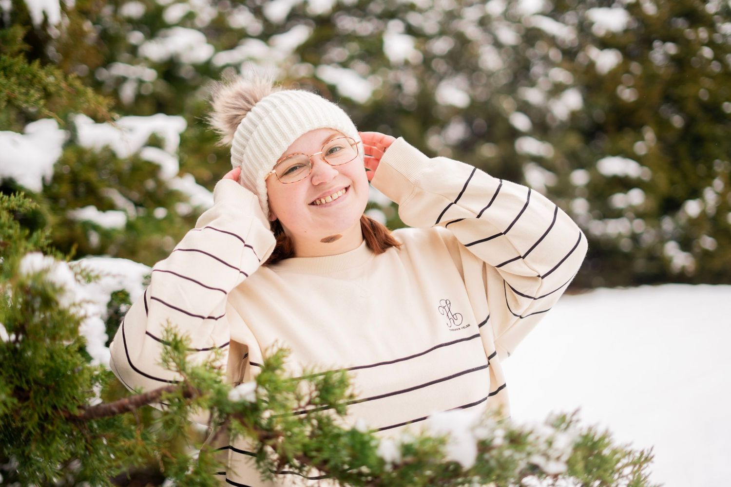 Young woman in striped sweater and white pom beanie smiles among snow-covered evergreen trees.