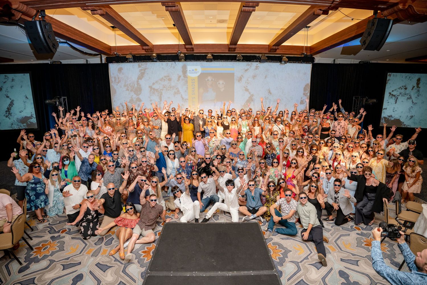 Large crowd of conference attendees pose for group photo in hotel ballroom with raised arms.