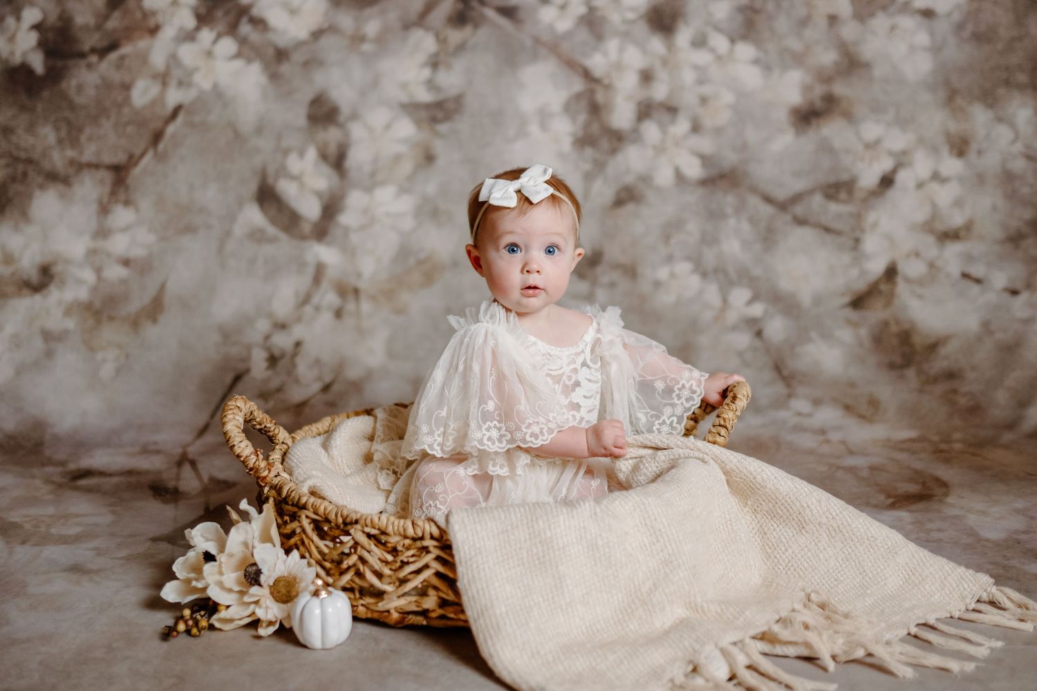 A vintage-style portrait of a baby sitting with toys in a cream-colored dress against a textured backdrop.