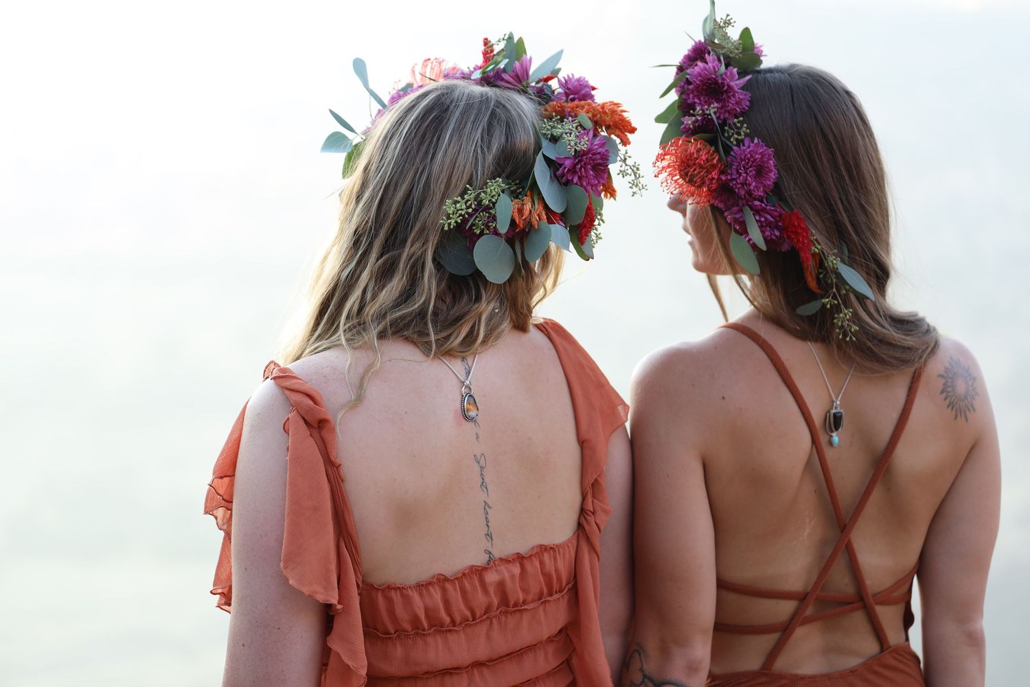 Two women wearing floral crowns and summer dresses against light background.