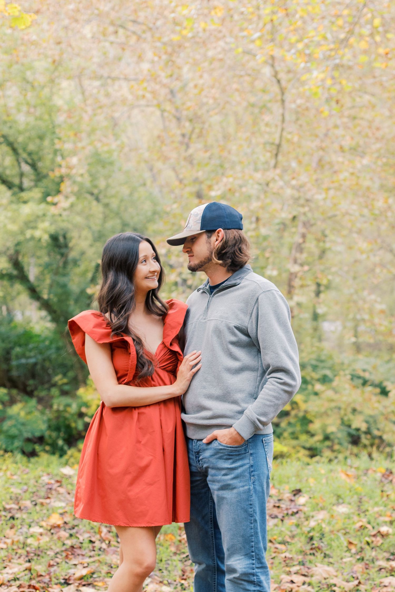 A couple shares a romantic moment in autumn, with her in a red dress and him in casual attire.