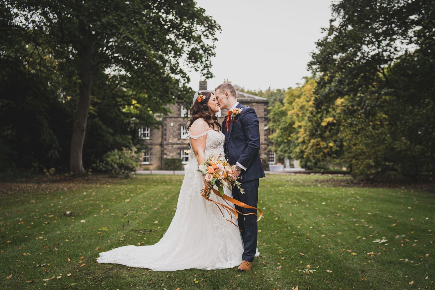 A romantic wedding couple embraces on a green lawn with an elegant manor house visible in the background.