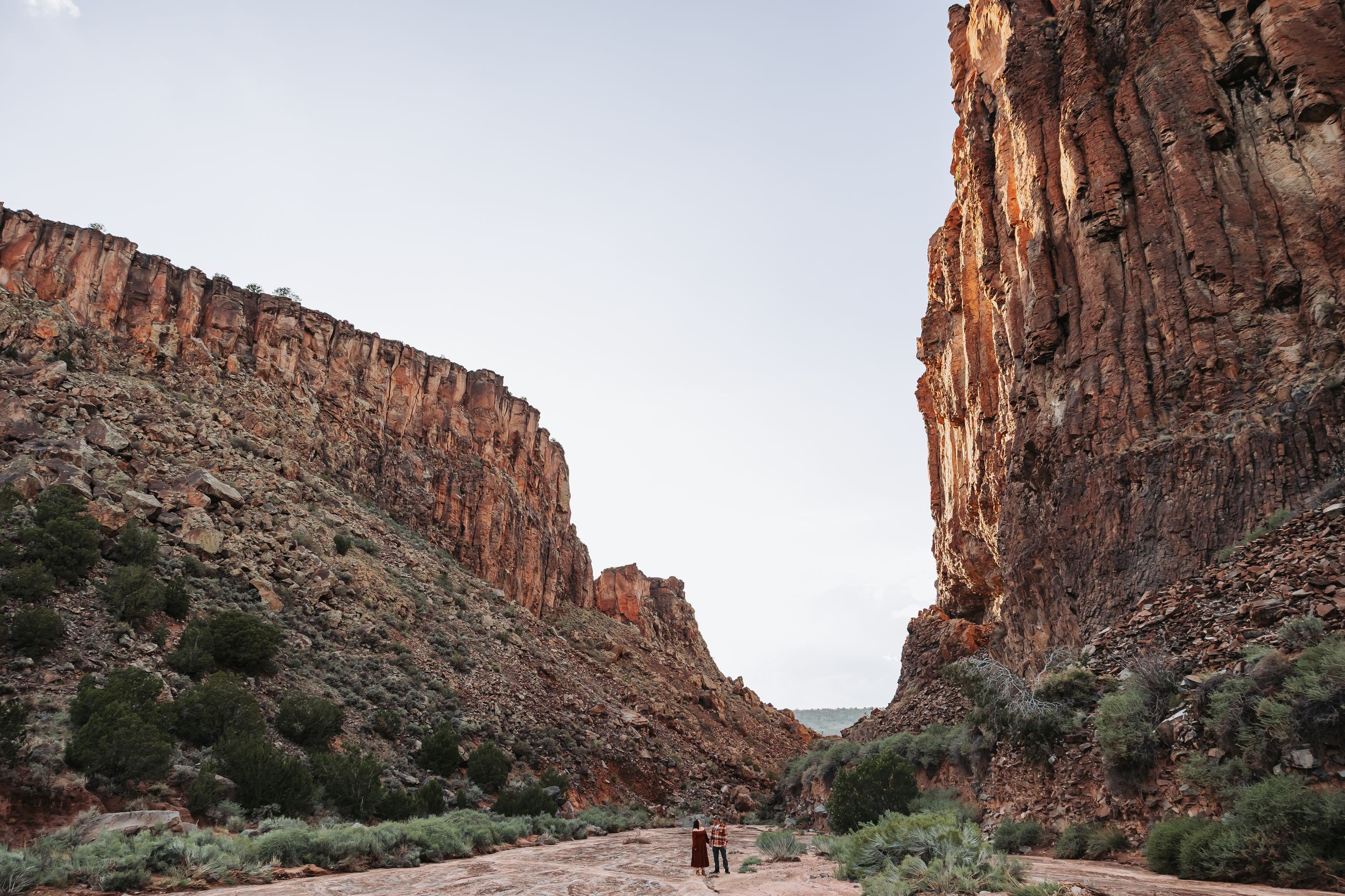 Two hikers walk through a dramatic red rock canyon with towering sandstone cliffs under a clear sky.