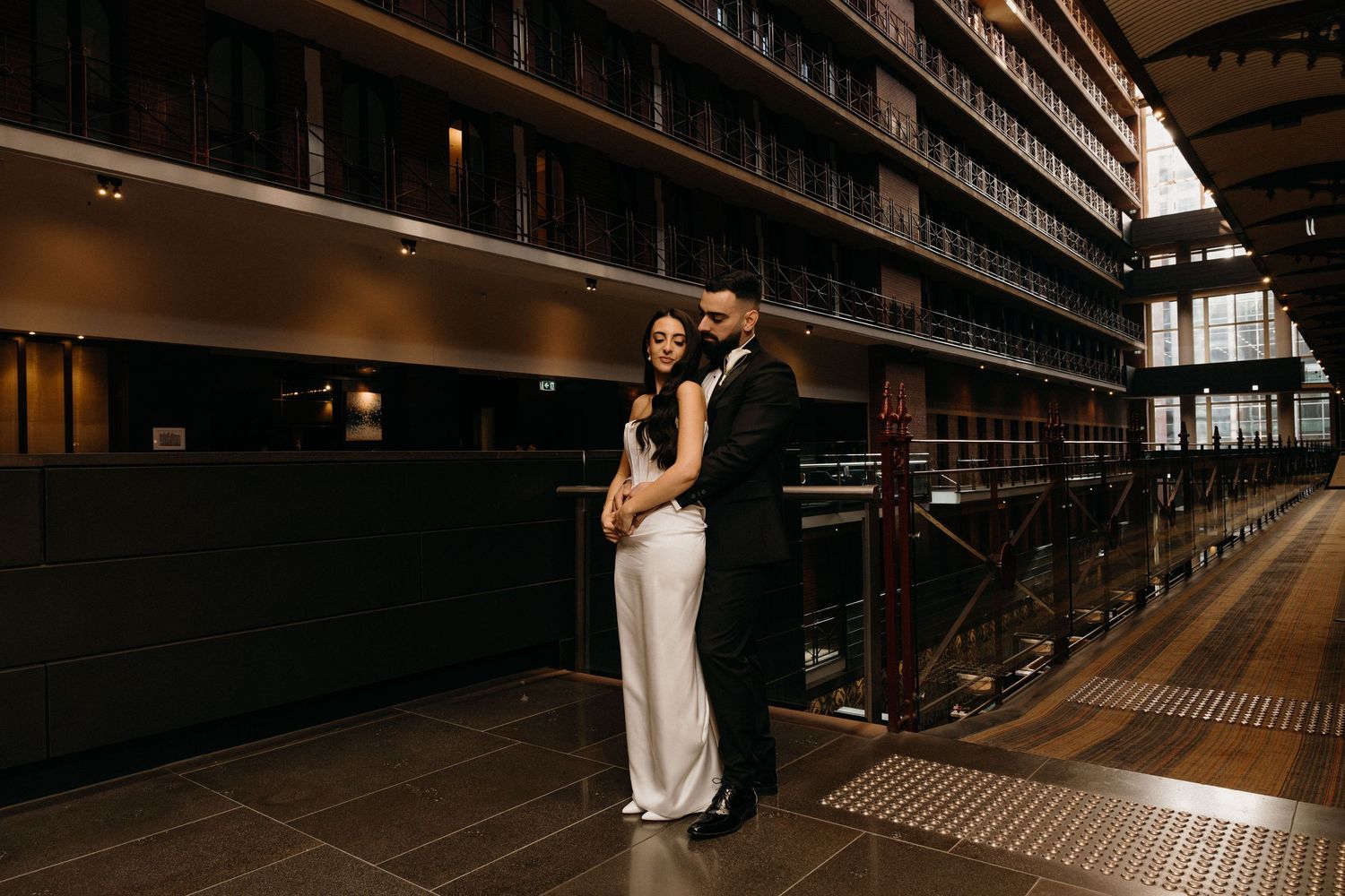Couple embracing in a grand library with tall bookshelves, wearing elegant wedding attire in a romantic pose.