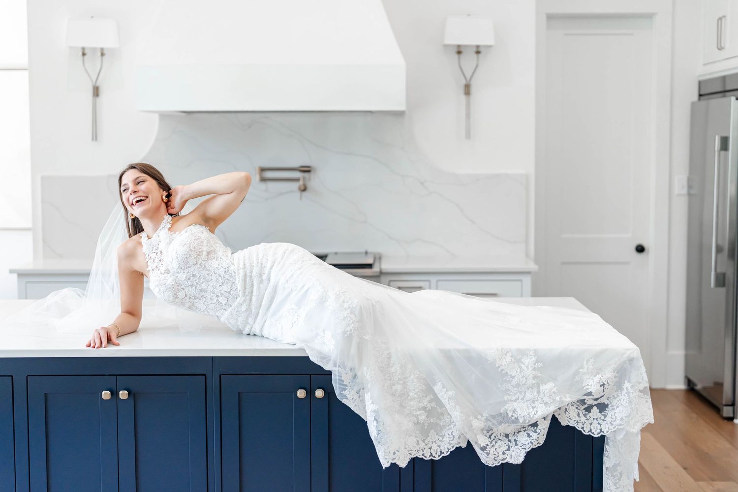 Bride in lace wedding gown posing elegantly on a navy blue kitchen island in a bright white luxury kitchen.