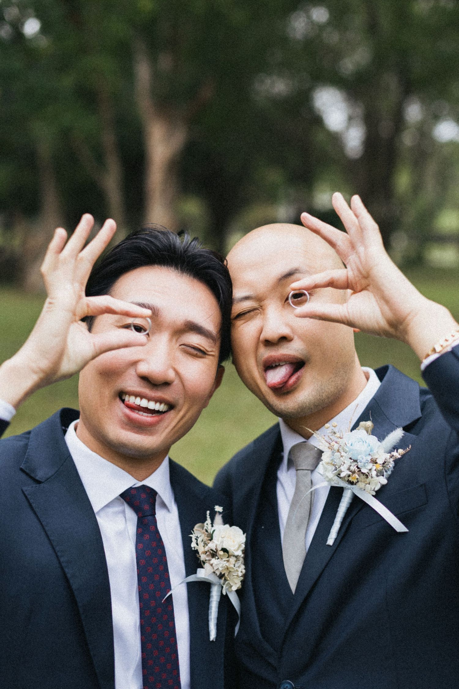 Two grooms in navy suits with boutonnieres make silly faces together outdoors on their wedding day.